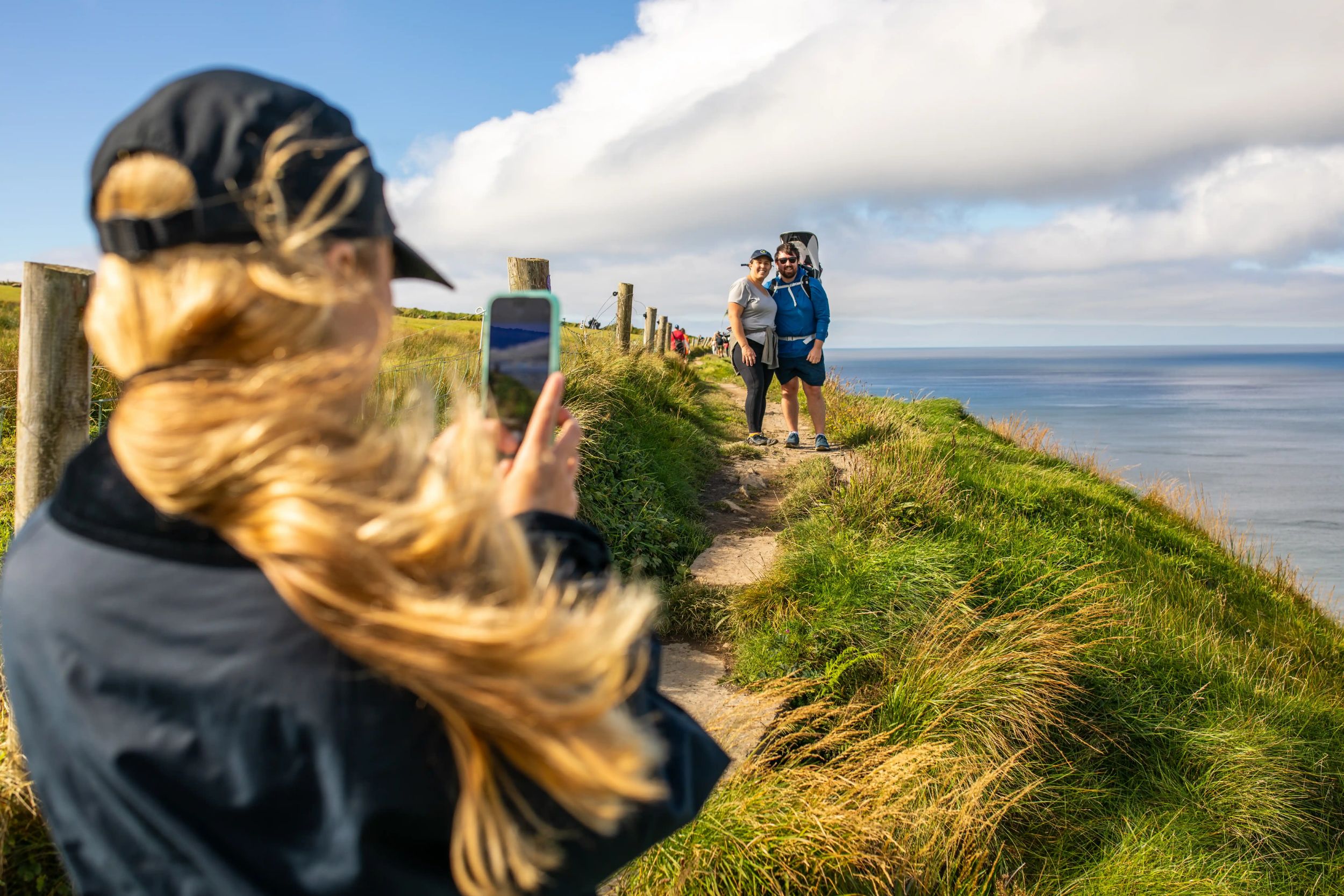 Woman photographing couple on scenic coastal path with sea views.