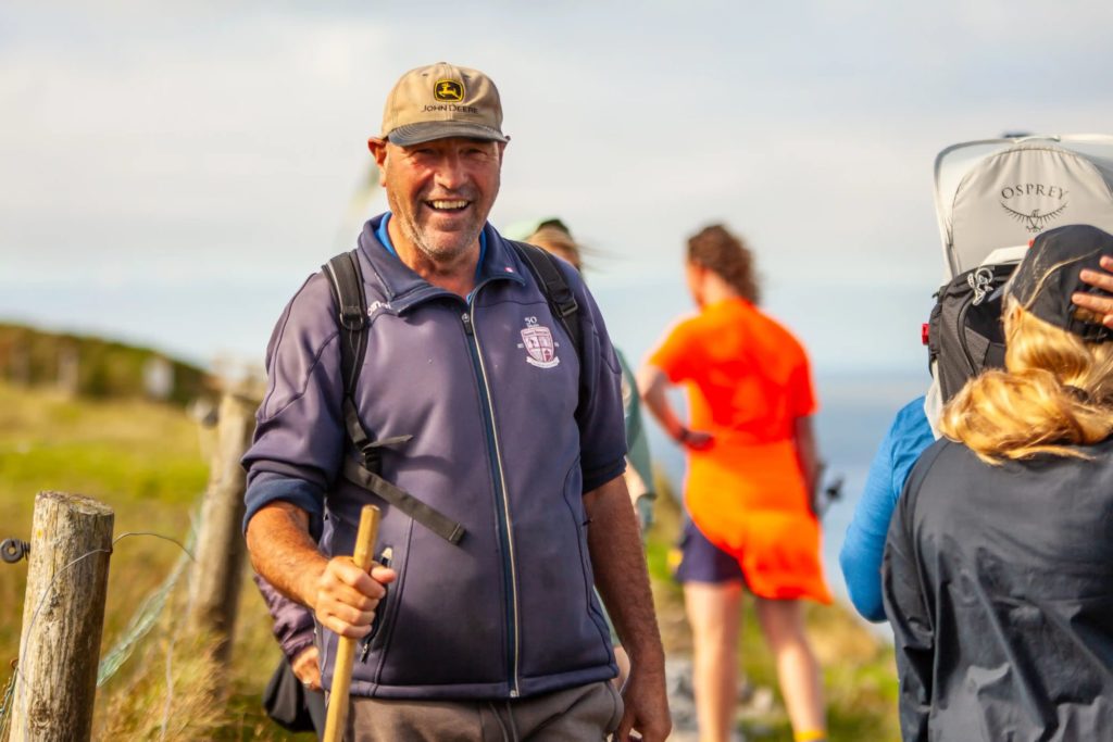 AKphoto (114) Man smiling while hiking with a group in a countryside setting, carrying a walking stick.