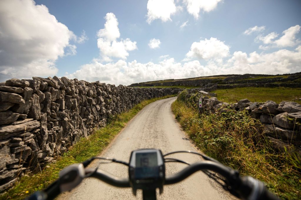AKphoto (118) Cyclist view along a narrow country lane surrounded by stone walls under a bright blue sky with fluffy clouds.
