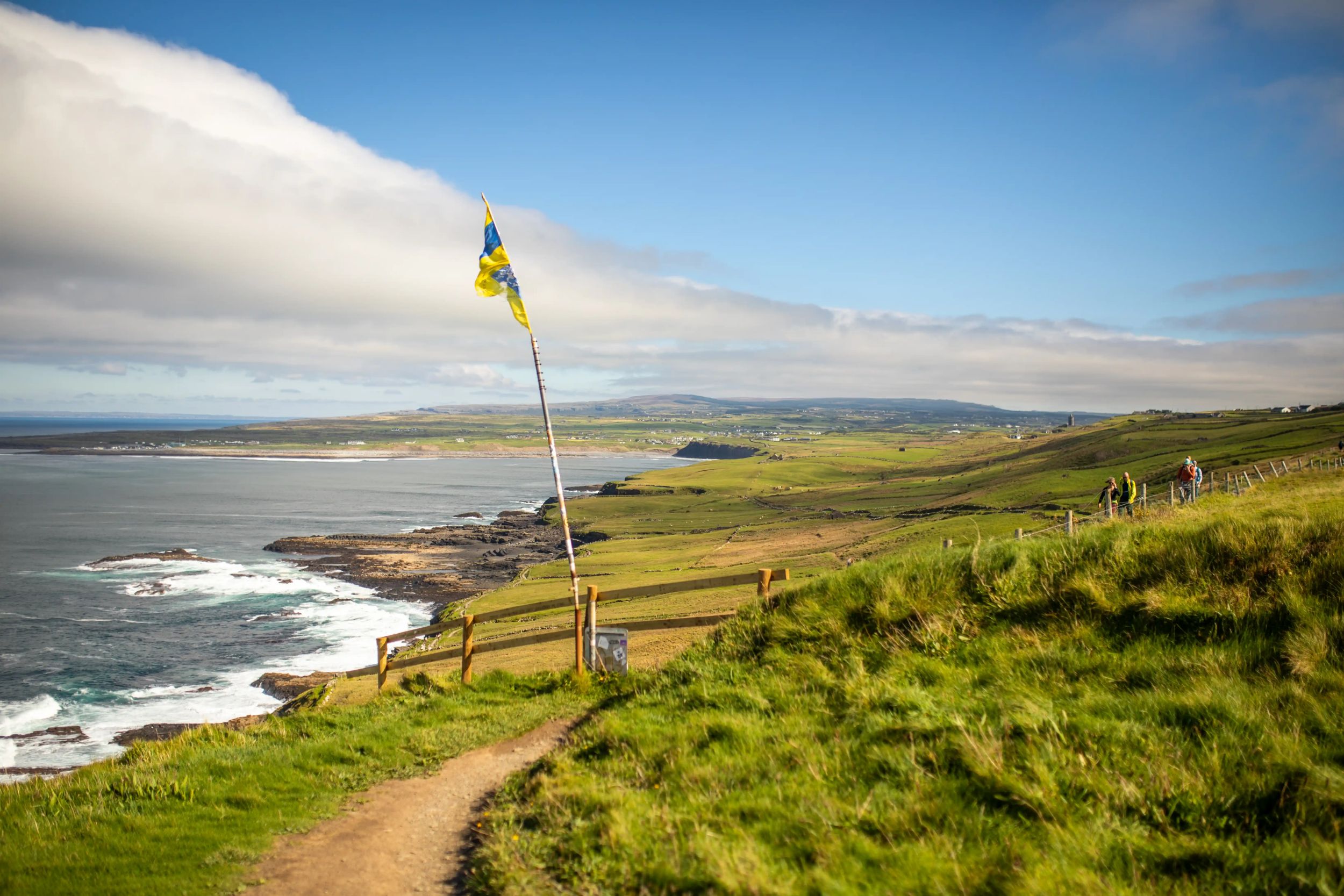 Scenic coastal view of Irish countryside with green hills, ocean cliffs, and hikers exploring a winding path under blue skies.