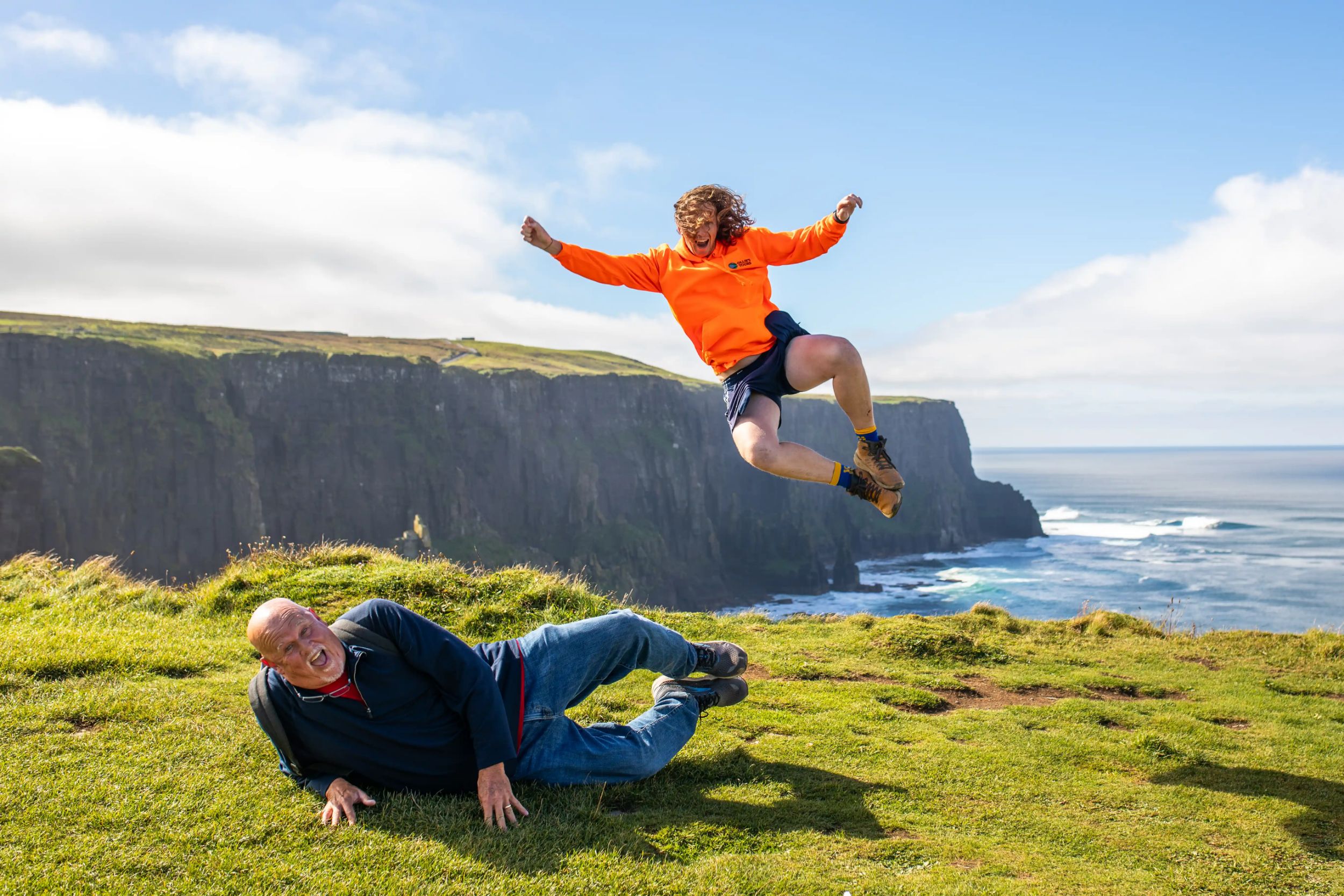 Two people having fun at the Cliffs of Moher: one jumping, one lying down, with scenic ocean views.