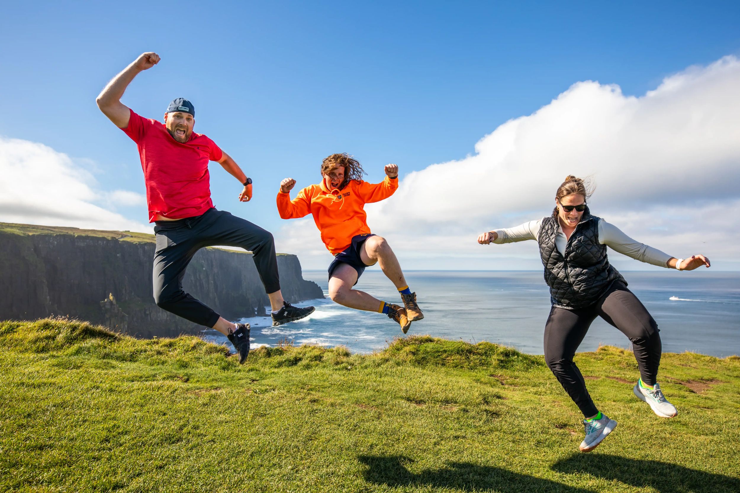 People joyfully jumping on a cliff edge with ocean view, sunny skies, and cliffs in the background.