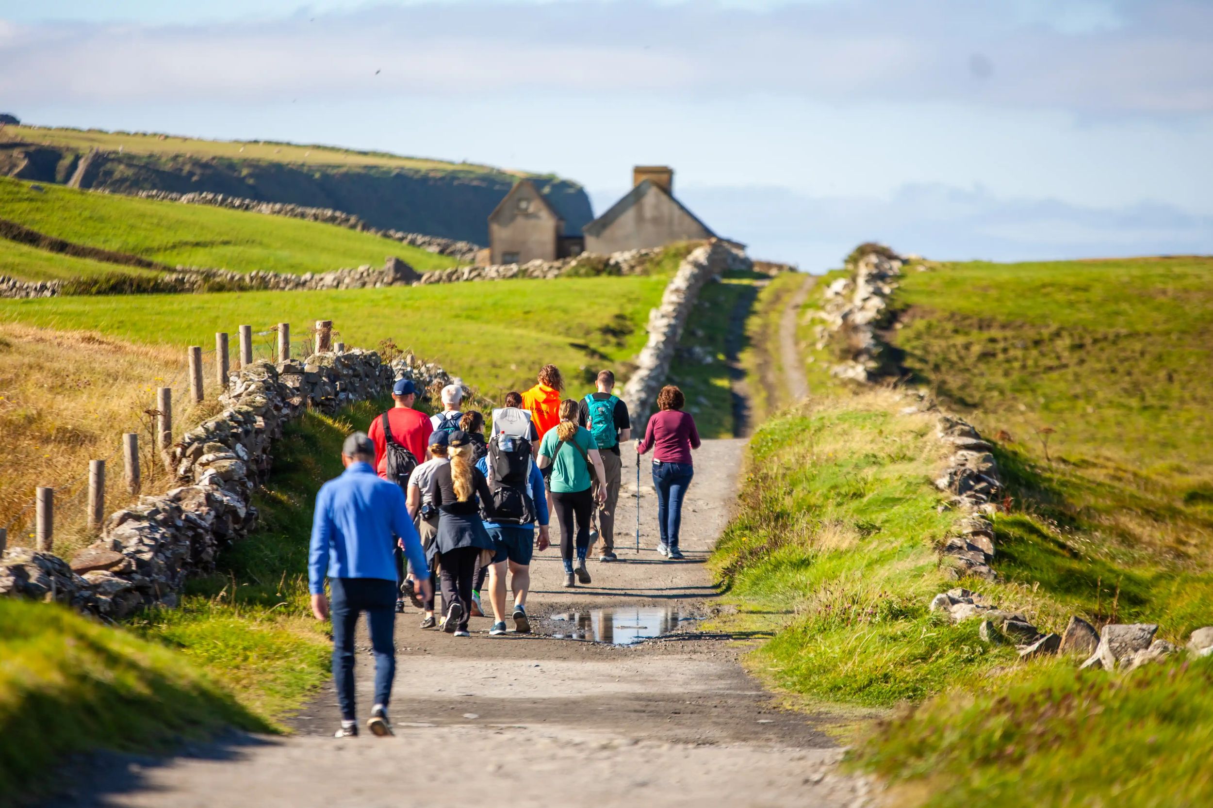 Group hiking along a scenic rural path with stone walls and green fields under a clear blue sky.