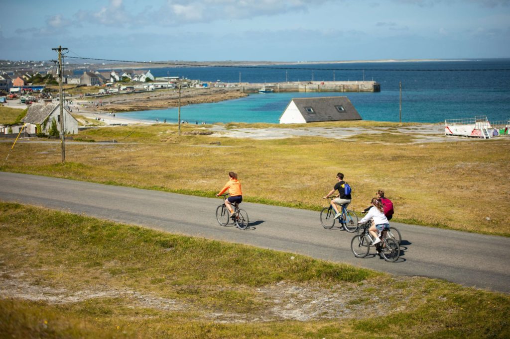 AKphoto (142) Cyclists ride along a scenic coastal road on a sunny day with a view of the sea and distant village.