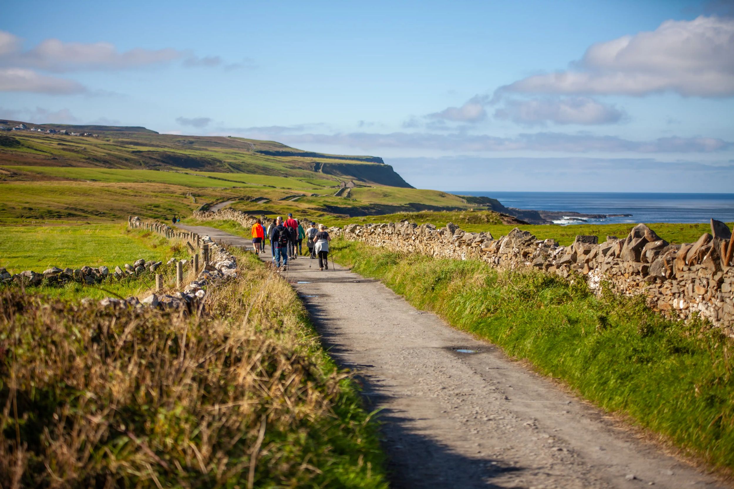 Group hiking along a coastal path with stone walls and lush green hills under a clear blue sky.
