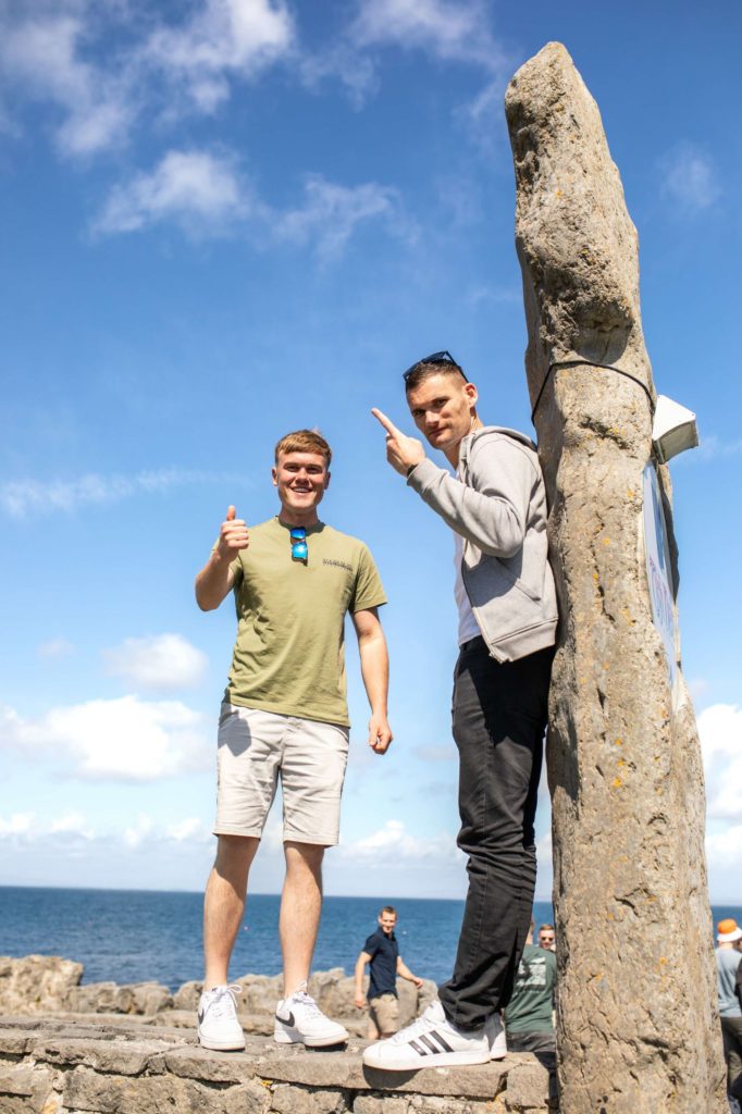 AKphoto (164) Two men posing beside a rock by the sea on a sunny day, giving thumbs up and pointing at the sky.