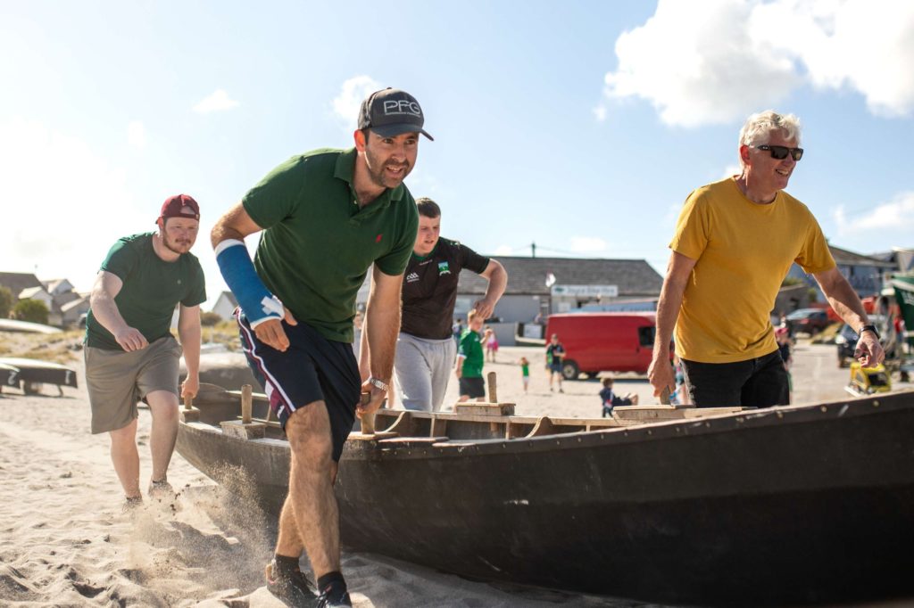 AKphoto (165) Group of men launching a traditional wooden boat on a sunny beach, showcasing teamwork and energy in Ireland.