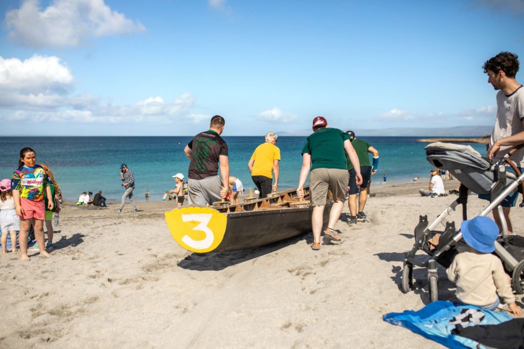 AKphoto (167) Group of people moving a boat on a sunny beach day, with children and families enjoying the seaside.