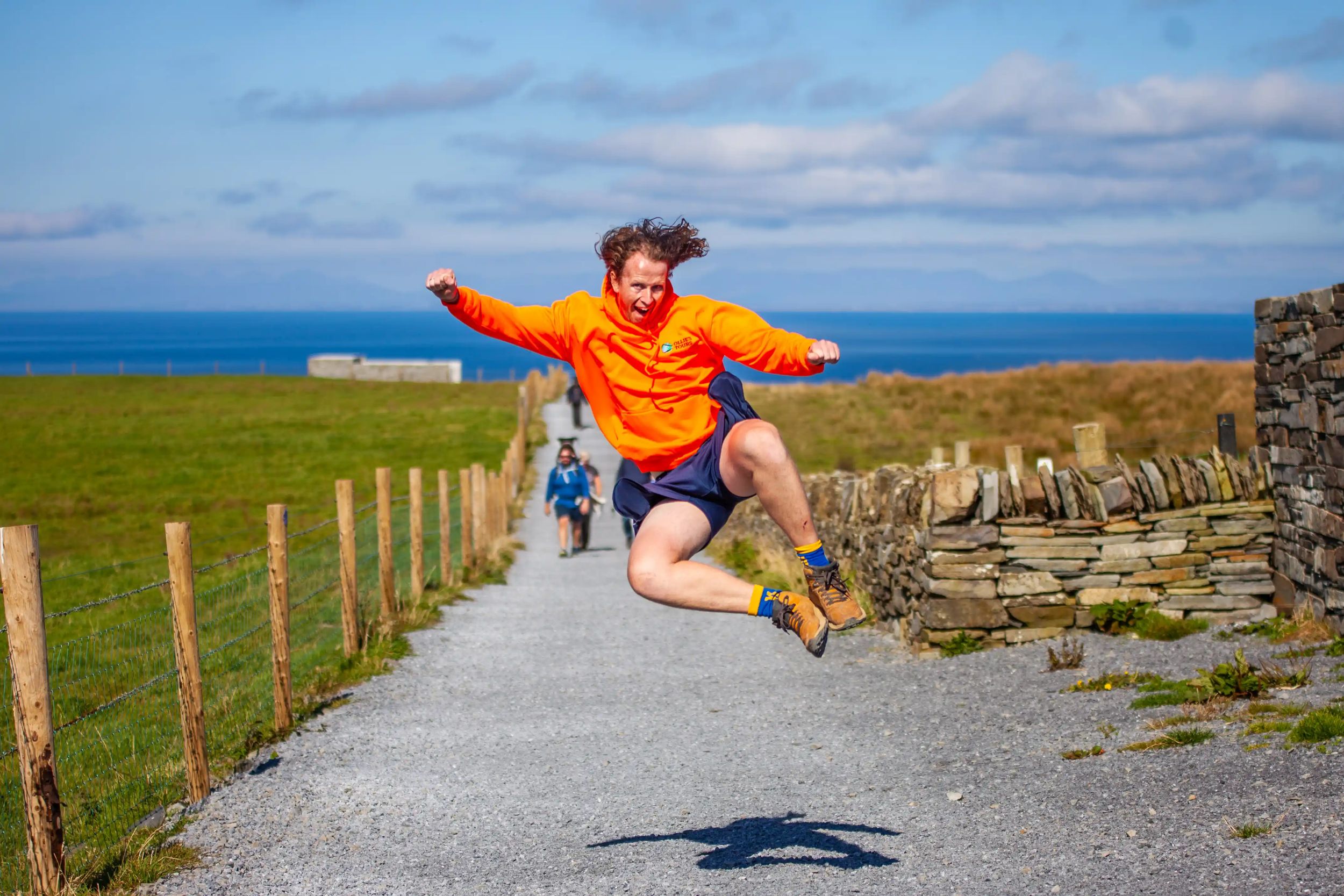 Ollie joyfully jumps on path by seaside, wearing an orange hoodie and hiking boots, with ocean view background.