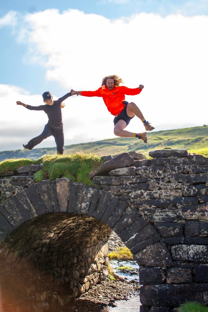 AKphoto (35) Two people energetically jumping on a stone bridge against a bright sky and lush landscape backdrop.