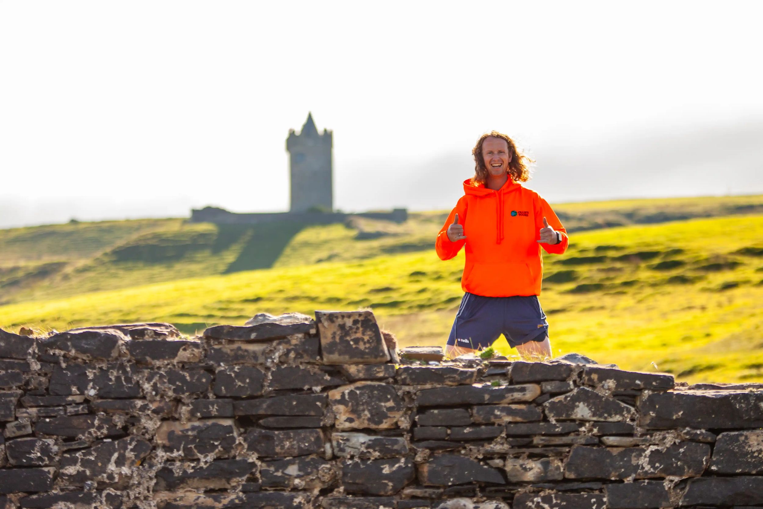 Ollie posing with thumbs up in front of Irish countryside and medieval tower.