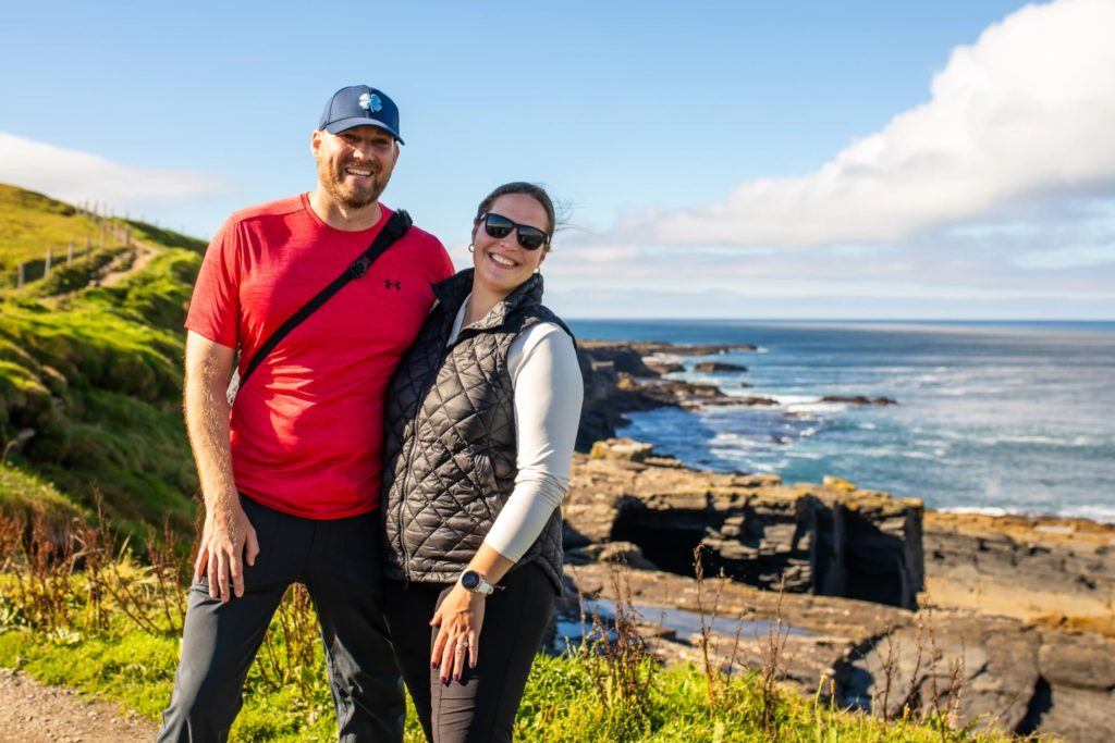 AKphoto (48) Couple smiling on a sunny coastal walk with cliffs and ocean view in the background.