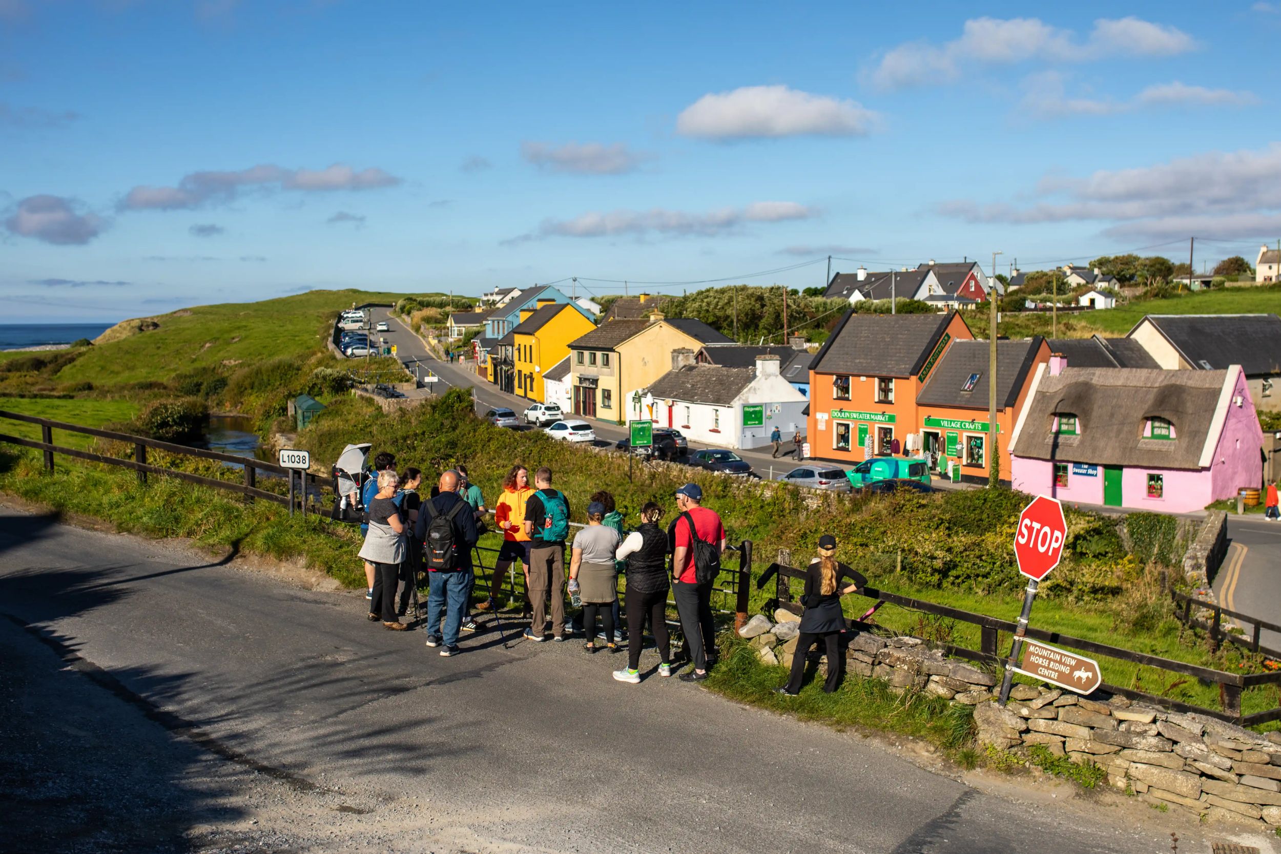 Group of tourists exploring a colorful Irish village on a sunny day, with green hills and ocean in the background.