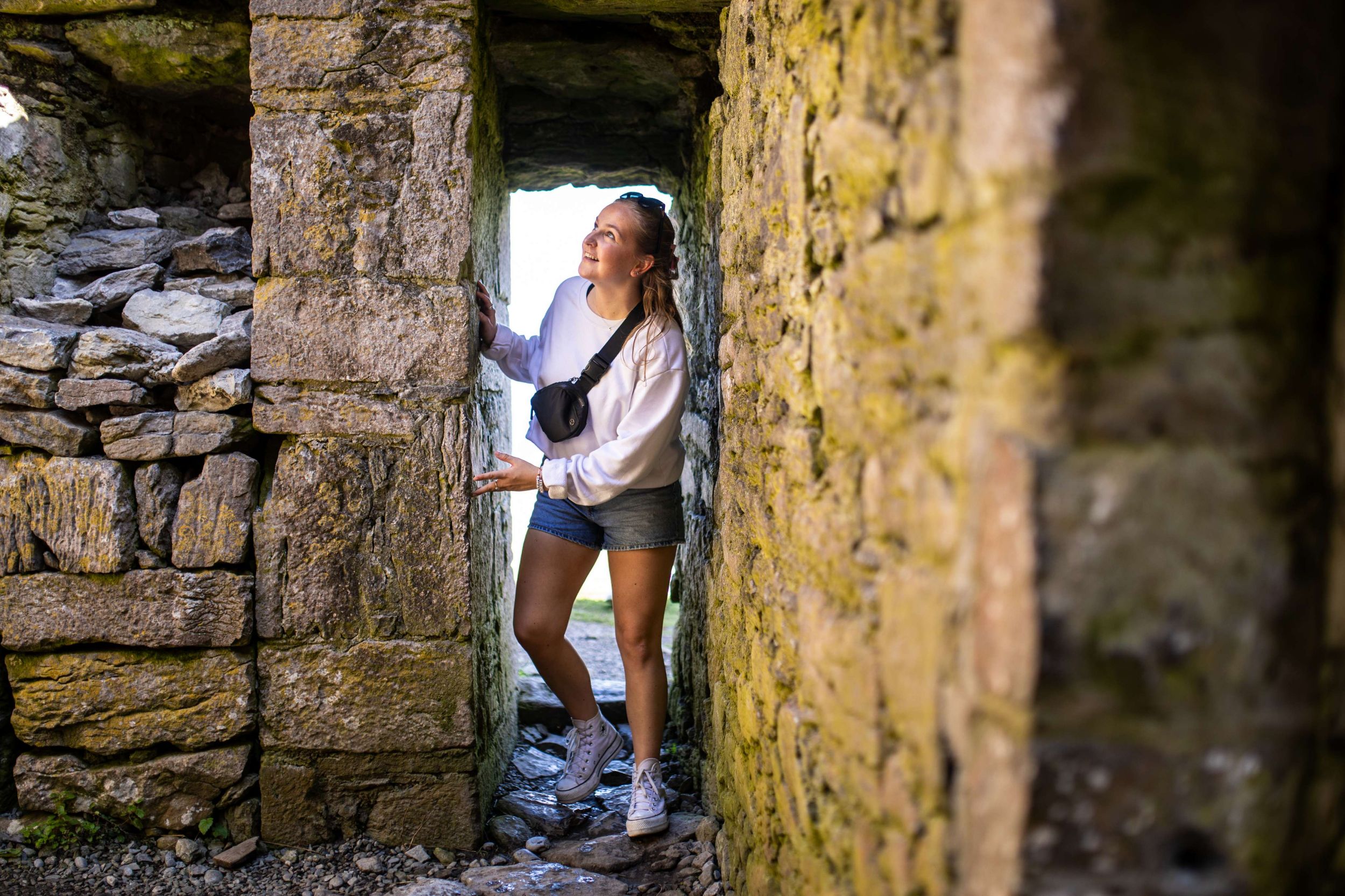Woman exploring a narrow stone passage in an ancient ruin, wearing casual clothes and smiling at the light ahead.