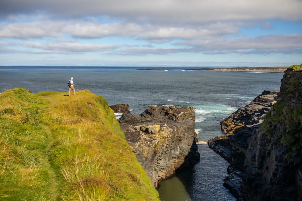 AKphoto (80) Hiker on stunning Irish coastal cliff path, overlooking ocean and rugged terrain under partly cloudy blue sky.
