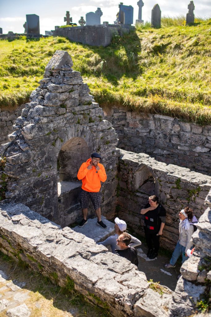 AKphoto (81) A group exploring ancient stone ruins and a graveyard under bright sunshine, guided by a person in an orange jacket.