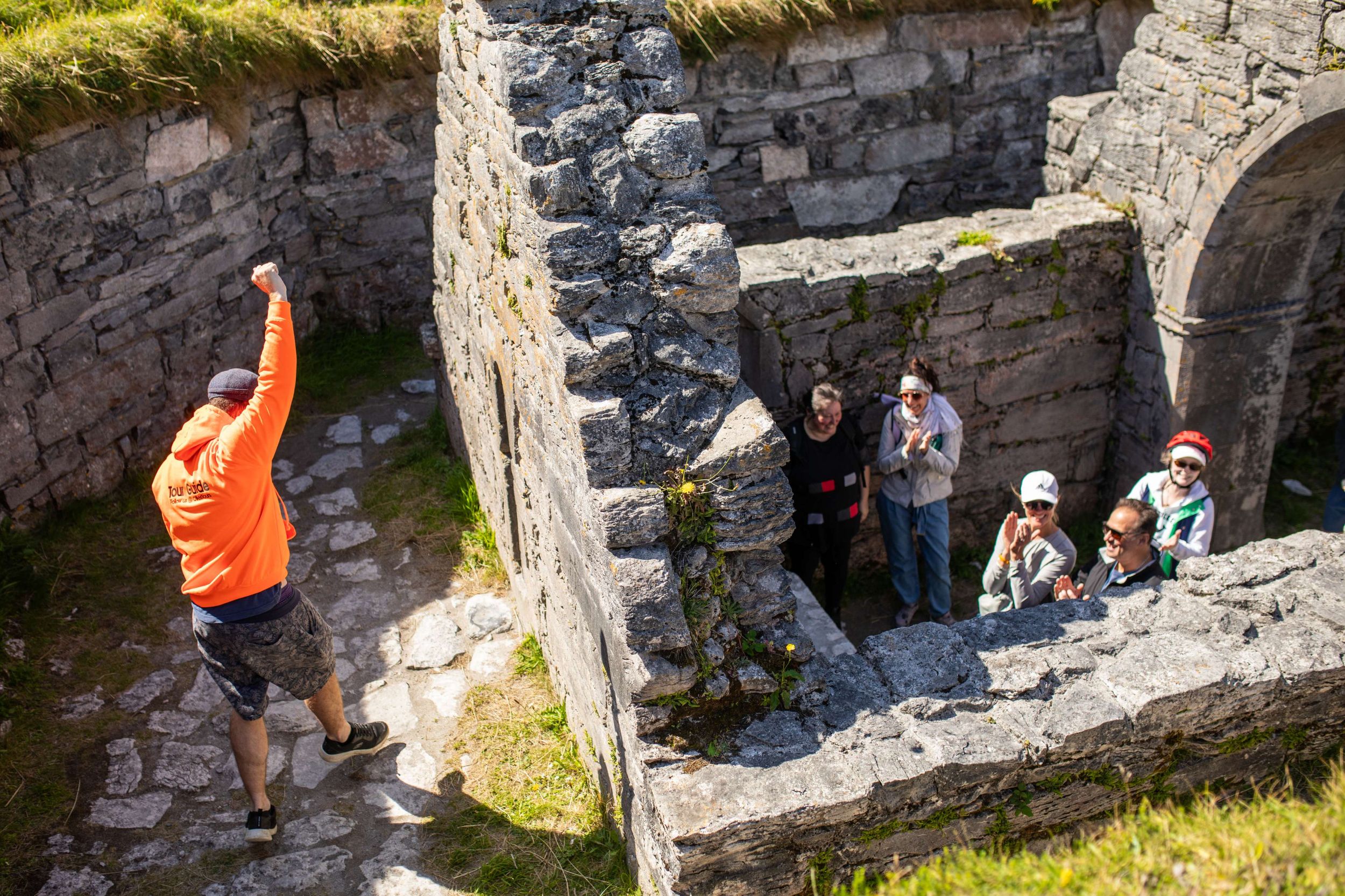 Ollie leading a group through ancient stone ruins on a sunny day, with visitors clapping and enjoying the experience.