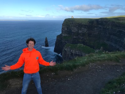 a man standing next to a body of water with Cliffs of Moher in the background
