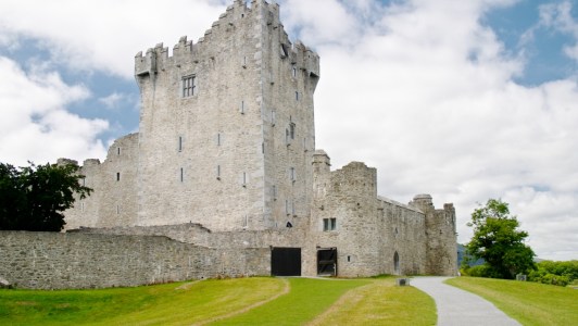 a stone castle with a clock tower in front of a brick building with Ross Castle in the background